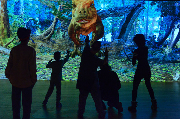 Shadows of people posing in front of a large T. rex dinosaur on a projection wall.