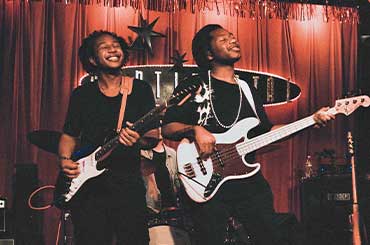 two Black men playing guitars and smiling on stage