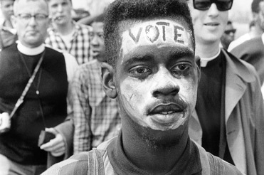 black and white close up photo of a man with the word VOTE painted on his forehead