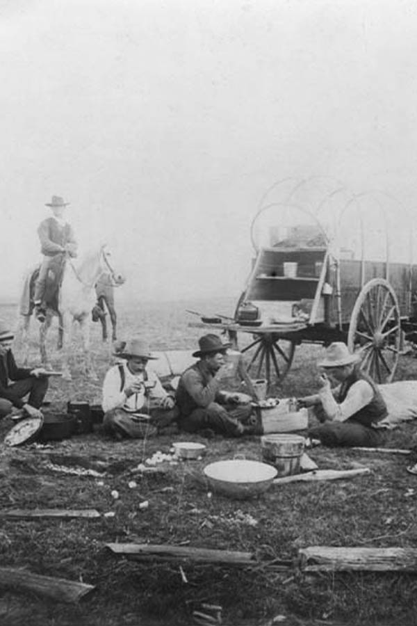 A sepia/black and white photo of a group of cowboys sitting on the ground with a wagon and a man on a horse behind them.