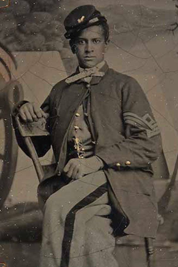 A sepia/black and white photo of a Black civil war soldier wearing a uniform and posing with a weapon.